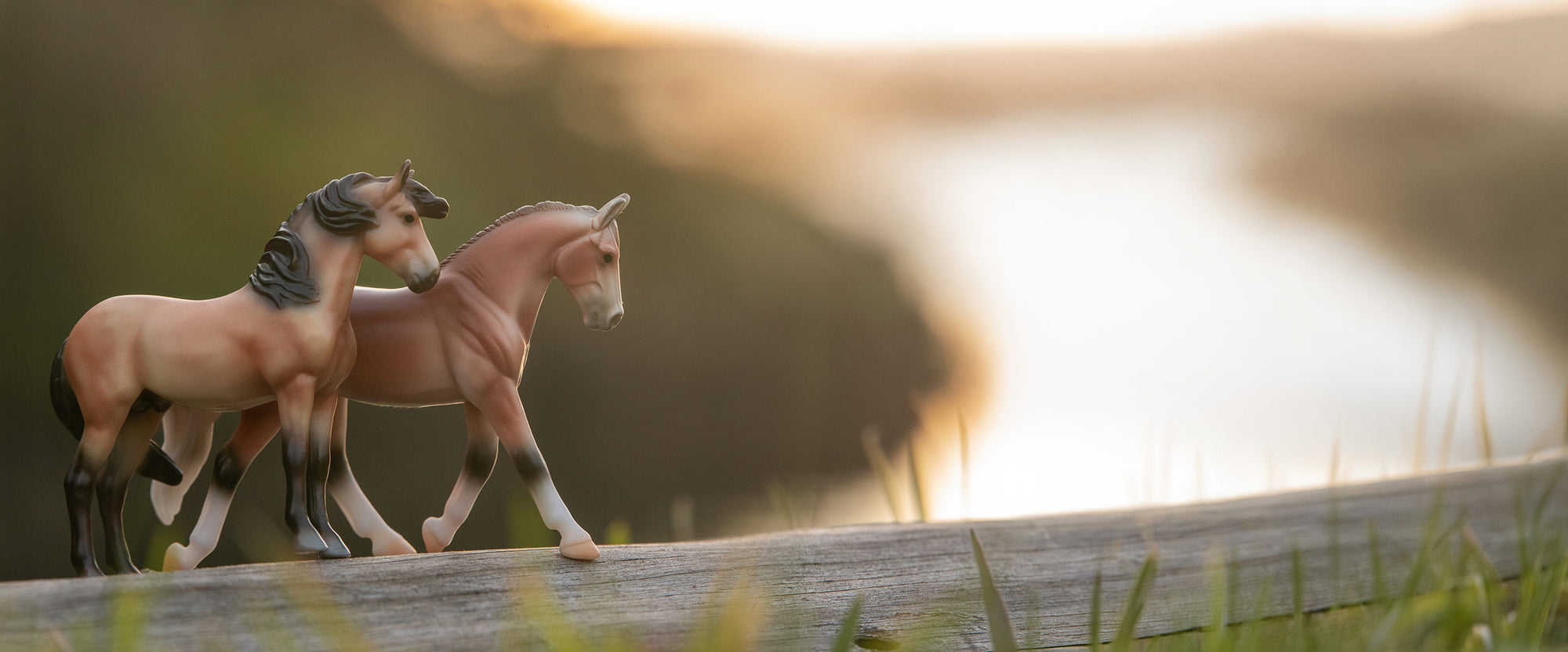 Two Breyer Stablemates standing side-by-side on a log overlooking a river
