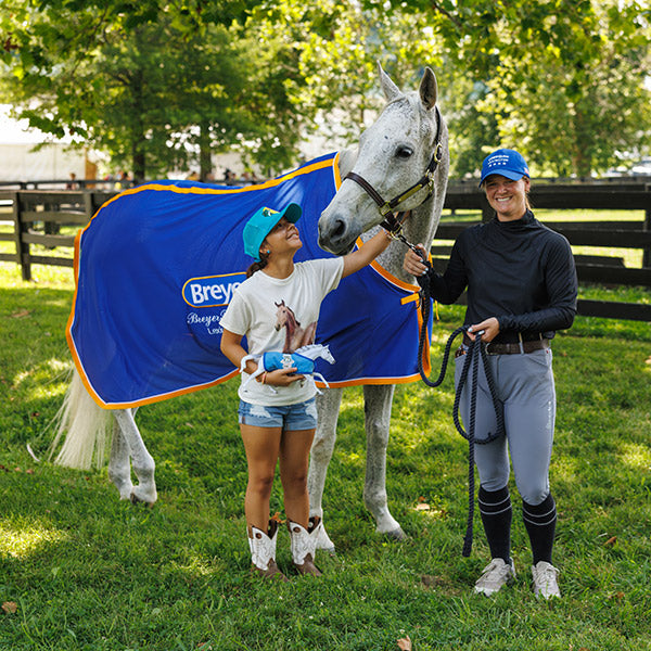 A child at BreyerFest holding a model and petting a live horse in a field with the horse's handler.