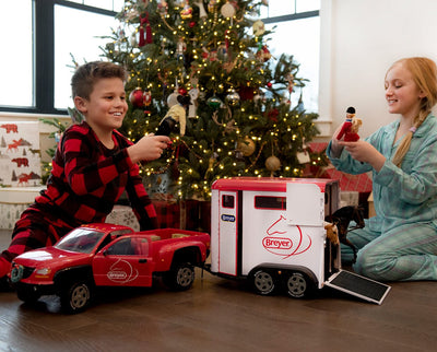 A girl and boy by the Christmas Tree playing with Benda, Austin and the Dually Truck and Trailer