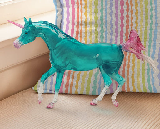 Sapphira | Unicorn facing left in front of a rainbow colored pillow. Translucent turquoise with pink horn, tail and hooves.