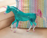 Sapphira | Unicorn facing left in front of a rainbow colored pillow. Translucent turquoise with pink horn, tail and hooves.